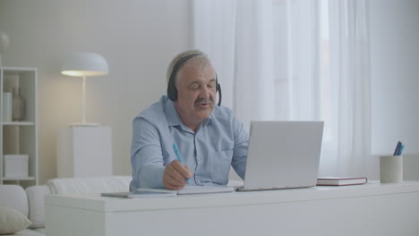 Un-Hombre-De-Mediana-Edad-Se-Comunica-Por-Internet-Usando-Una-Computadora-Portátil-Y-Auriculares-Tomando-Y-Tomando-Notas-En-Un-Cuaderno